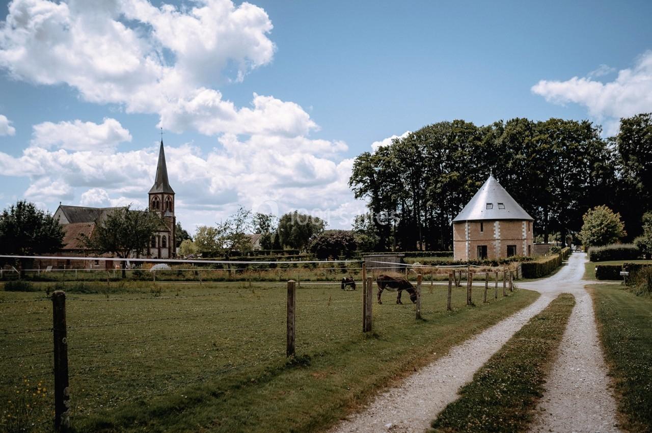 Chemin de campagne bordé de clôtures menant à une église et un bâtiment en pierre, sous un ciel partiellement nuageux.