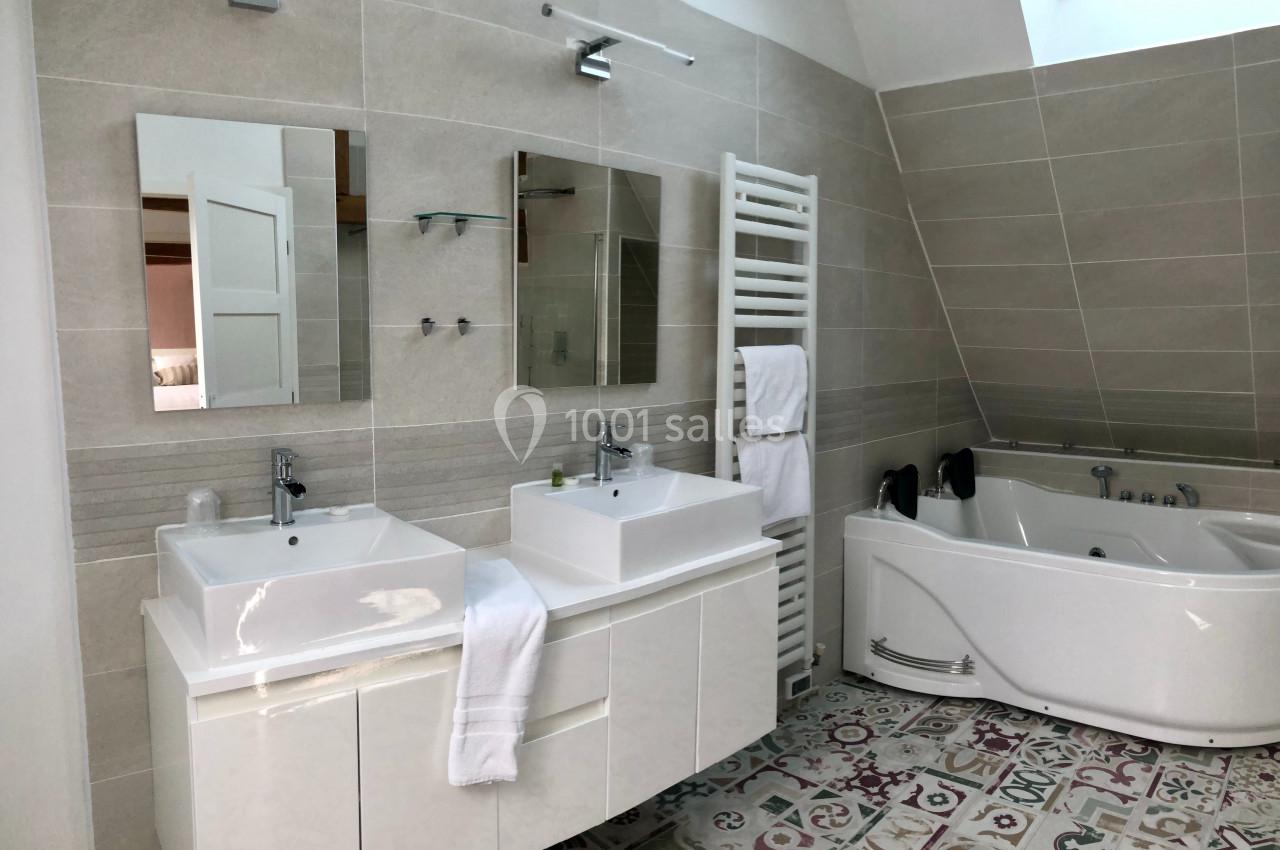 Location salle Argueil (Seine-Maritime) - Domaine de Clairval #27 Salle de bain moderne avec double vasque, baignoire d'angle, sèche-serviettes et carrelage décoratif au sol.