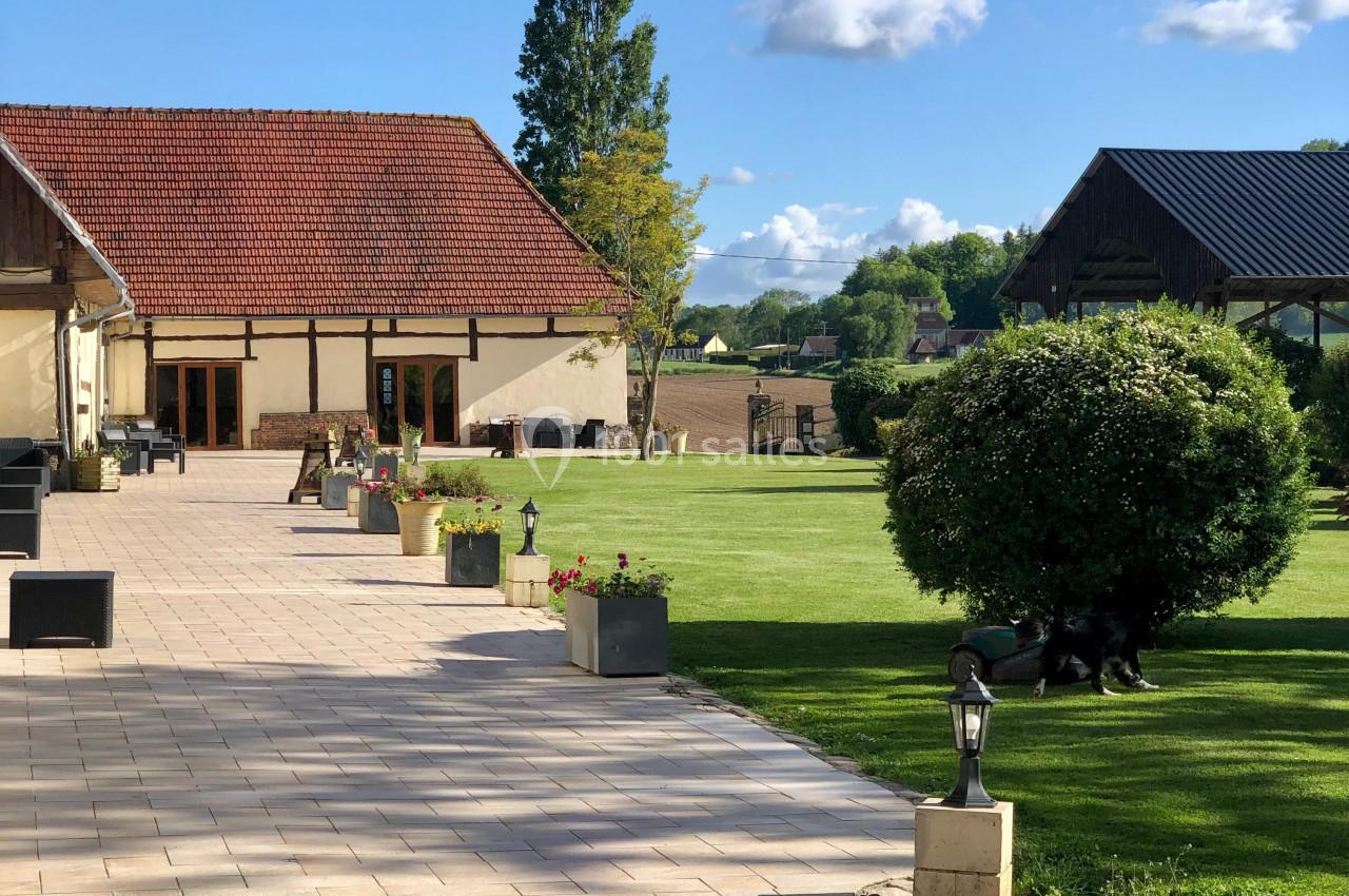 Location salle Argueil (Seine-Maritime) - Domaine de Clairval #2 Allée pavée bordée de fleurs menant à des bâtiments en pierre et à une pelouse arborée sous un ciel dégagé.