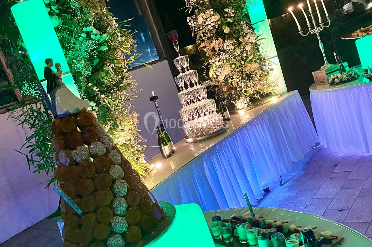 Location salle Argueil (Seine-Maritime) - Domaine de Clairval #22 Pièce montée éclairée en vert devant une pyramide de coupes et des décorations florales dans une salle de réception.