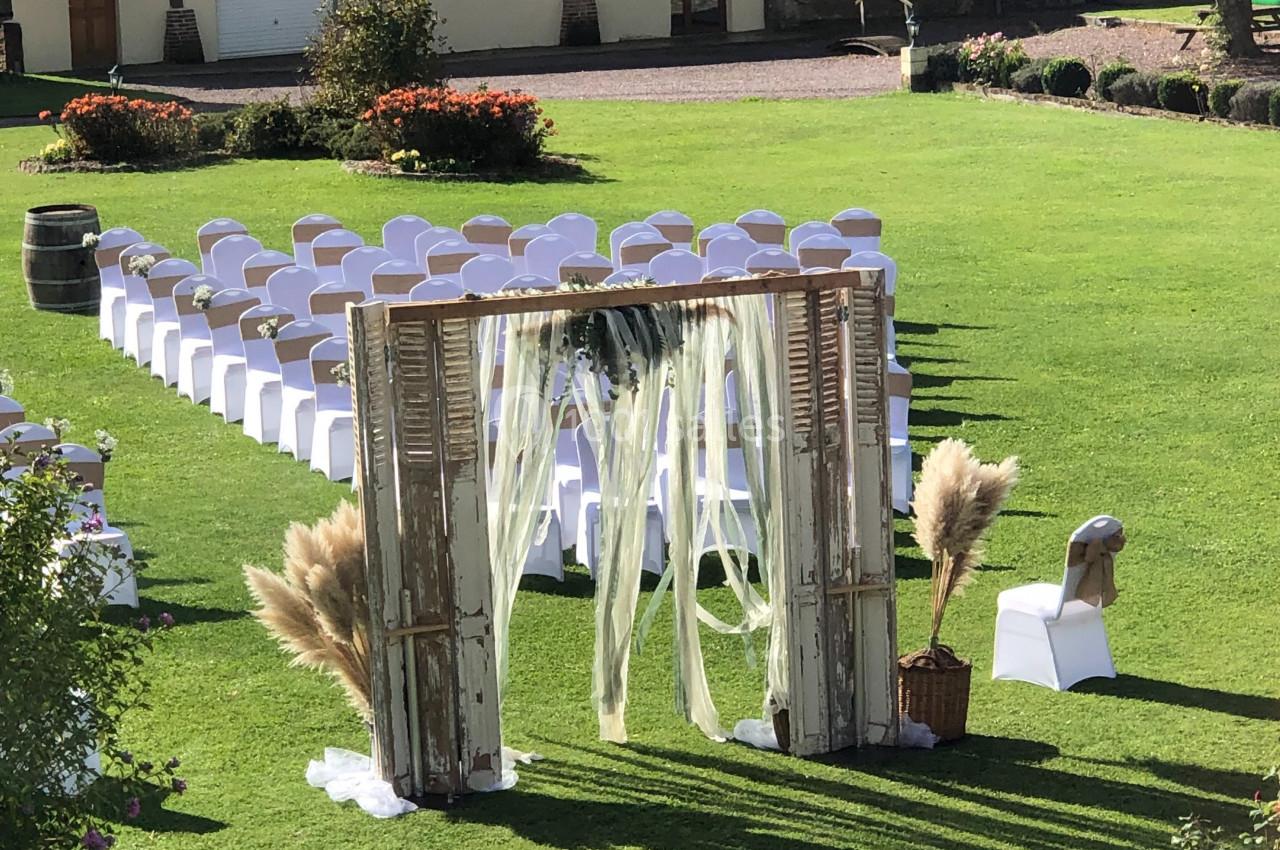 Location salle Argueil (Seine-Maritime) - Domaine de Clairval #15 Allée de chaises blanches alignées sur une pelouse, avec une arche décorée de voilages et de fleurs pour une cérémonie en…