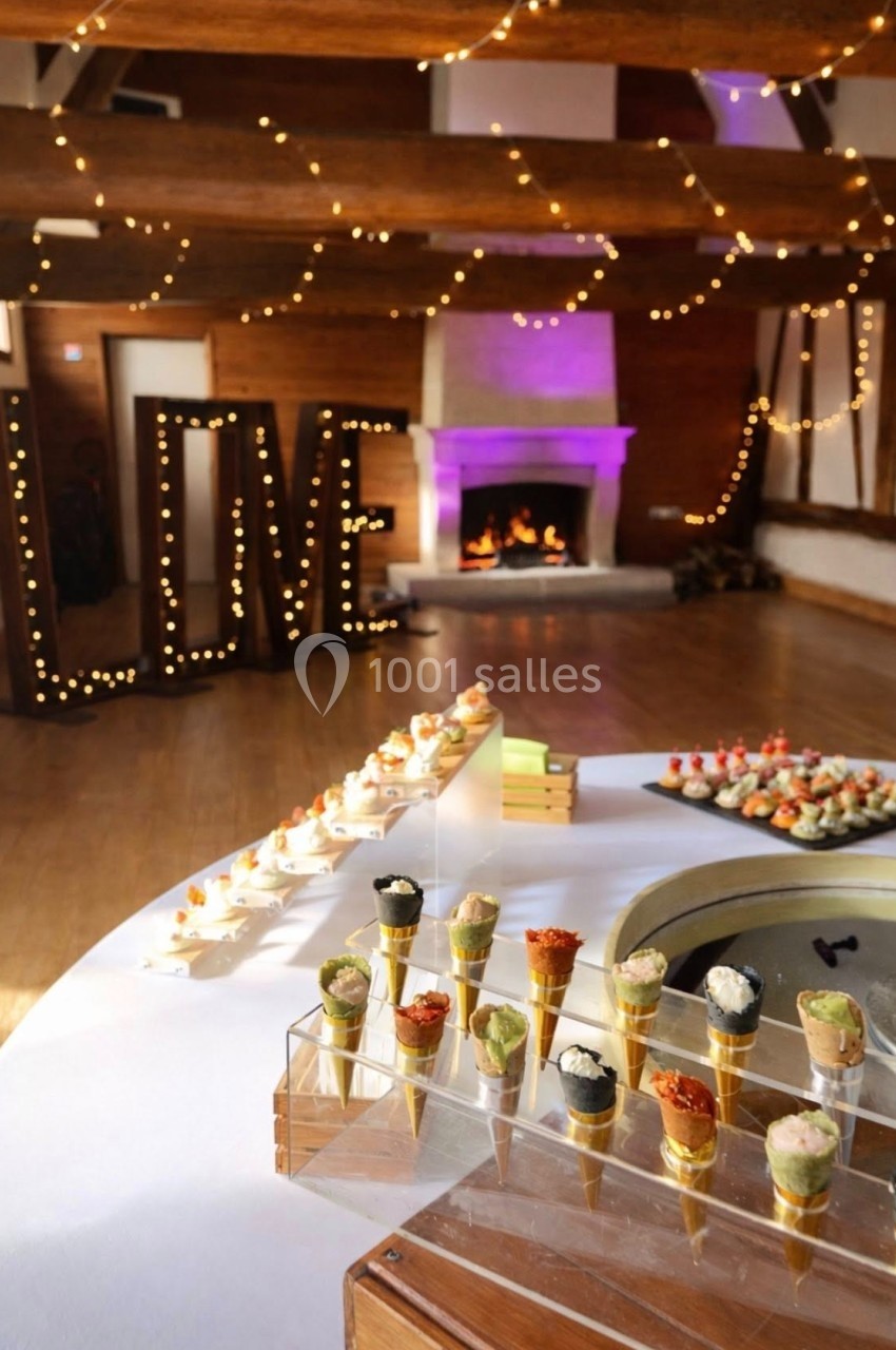 Location salle Argueil (Seine-Maritime) - Domaine de Clairval #2 Buffet de réception avec cônes apéritifs, cheminée allumée et décor lumineux formant le mot ’LOVE’ en arrière-plan.