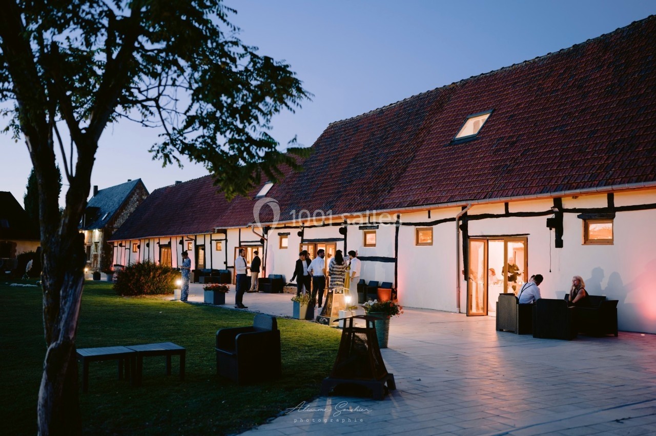 Location salle Argueil (Seine-Maritime) - Domaine de Clairval #5 Bâtiment à colombages éclairé en soirée, avec des personnes discutant sur une terrasse et un jardin à proximité.
