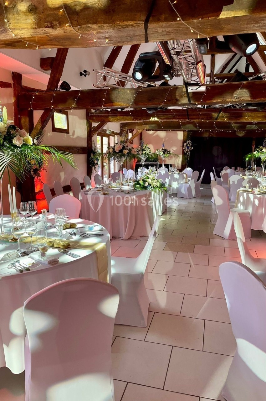 Location salle Argueil (Seine-Maritime) - Domaine de Clairval #11 Salle de réception décorée avec des tables rondes, nappes blanches, fleurs et éclairage chaleureux sous des poutres en bois.