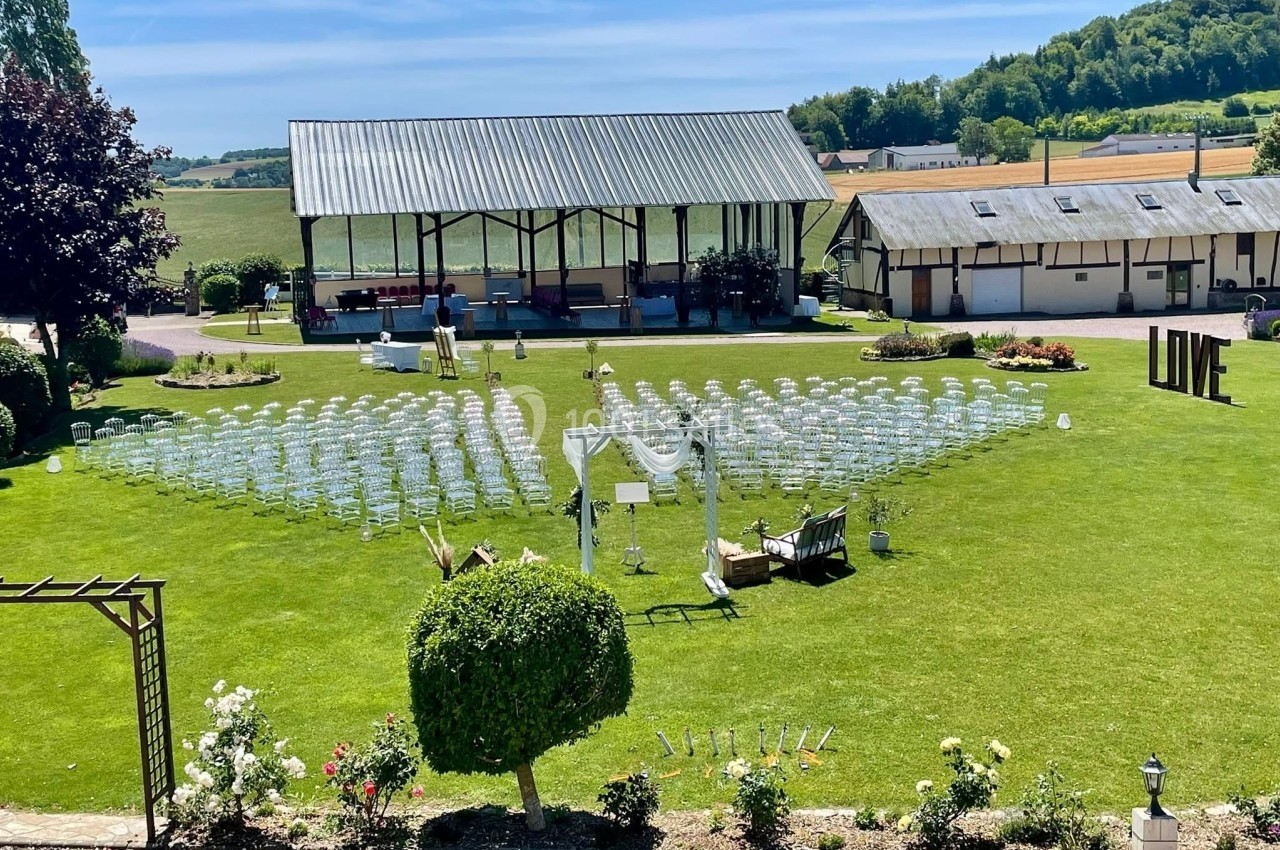 Location salle Argueil (Seine-Maritime) - Domaine de Clairval #15 Chaises disposées en extérieur sur une pelouse pour une cérémonie, avec une arche décorée et un bâtiment en arrière-plan.