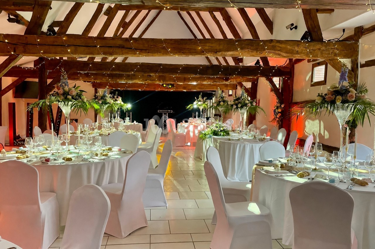 Salle de réception décorée avec des tables rondes dressées, nappes blanches et arrangements floraux, éclairage chaleureux.