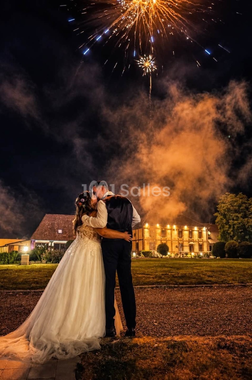 Un couple en tenue de mariage s'embrasse sous des feux d'artifice, devant un bâtiment éclairé de nuit.