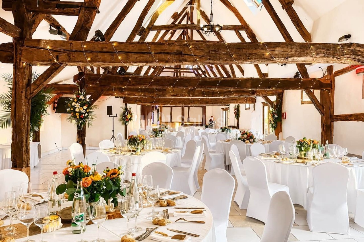 Salle de réception lumineuse avec poutres apparentes, tables rondes dressées et décorations florales élégantes.