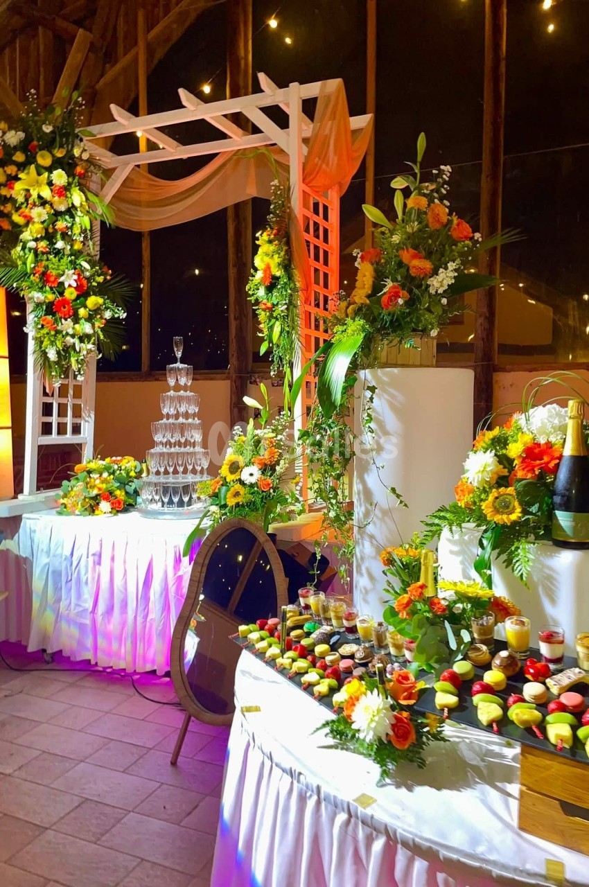 Buffet décoré avec des fleurs colorées, une pyramide de flûtes à champagne et des desserts variés sur des tables éclairées.