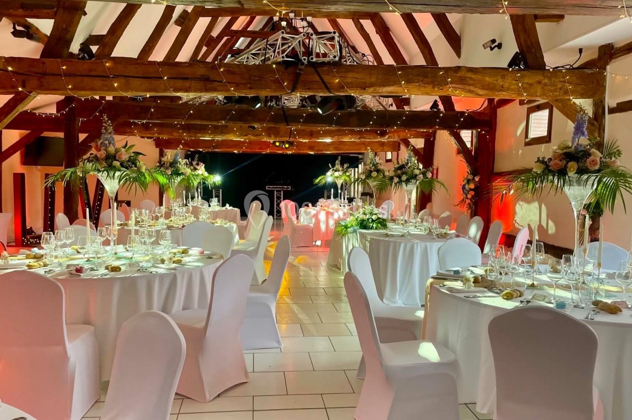 Salle de réception décorée avec des tables rondes dressées, chaises blanches et guirlandes lumineuses suspendues.