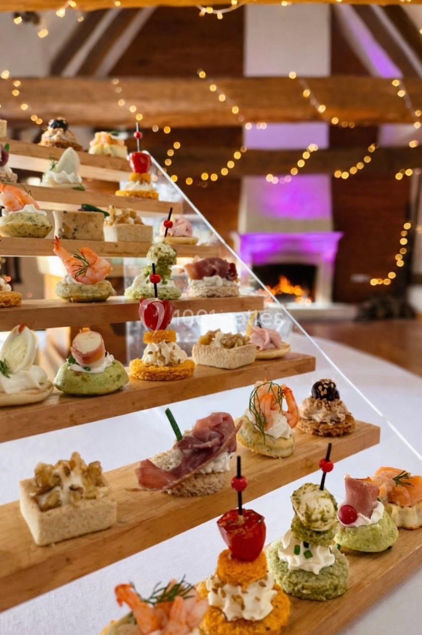 Cocktail - Grande Clairval (coté cheminée) Plateau en escalier avec divers amuse-bouches colorés dans une salle décorée de guirlandes lumineuses et cheminée allumée.