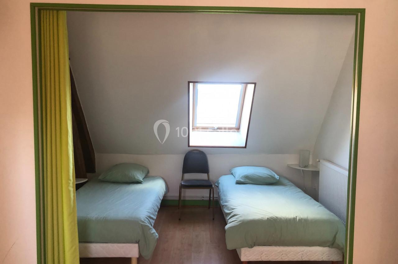 Dortoir BOX - Intérieur Deux lits simples avec draps verts dans une chambre mansardée, éclairée par une fenêtre de toit.