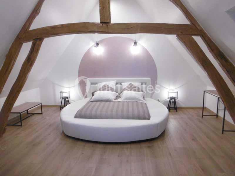 La petite maison dans la prairie - chambre des mariés Chambre mansardée avec lit rond, poutres apparentes, éclairage mural et mobilier minimaliste.
