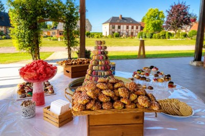 Miniature Vin d'honneur sous la Halle Table garnie de pâtisseries, macarons, fruits et biscuits, disposée en extérieur avec un jardin en arrière-plan.