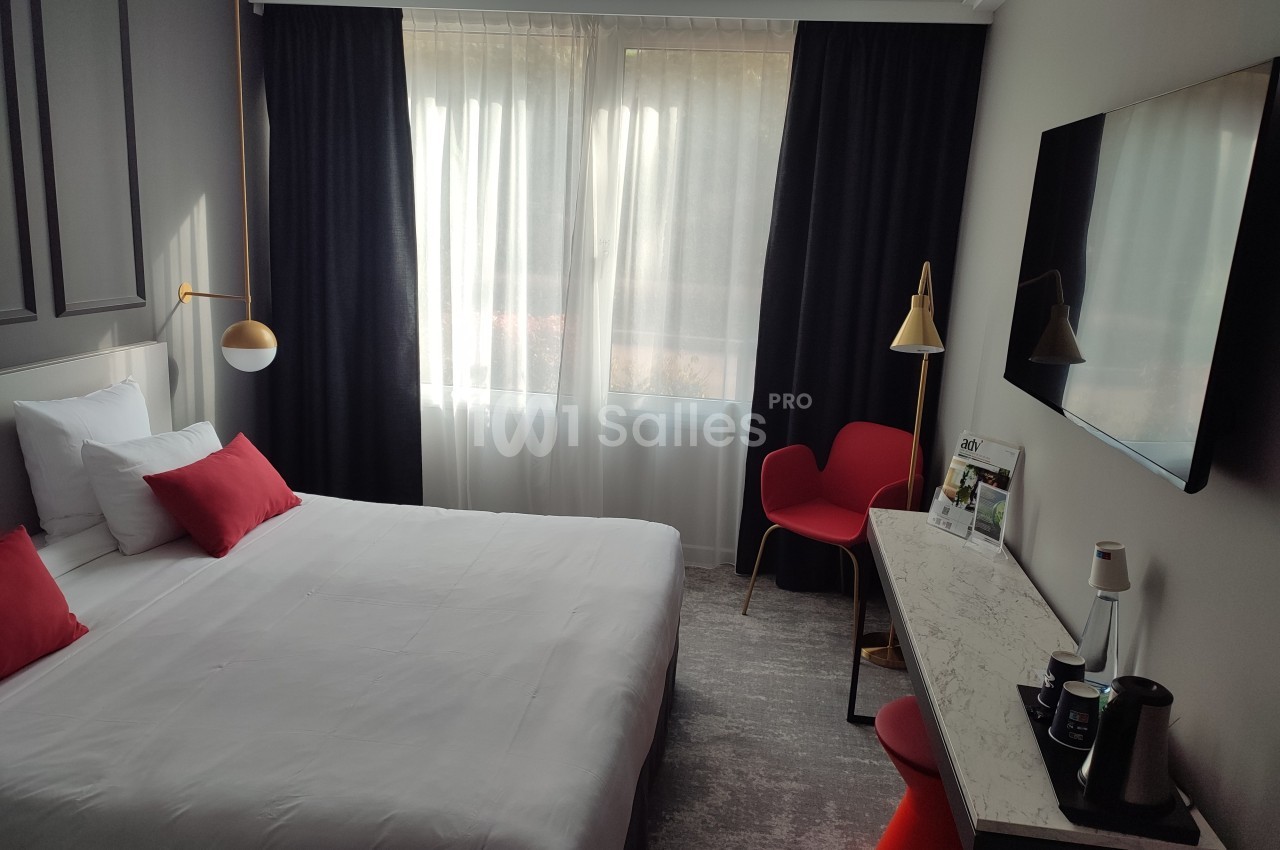 Chambre d'hôtel lumineuse avec un lit double, des coussins rouges, un bureau, une chaise et une télévision murale.