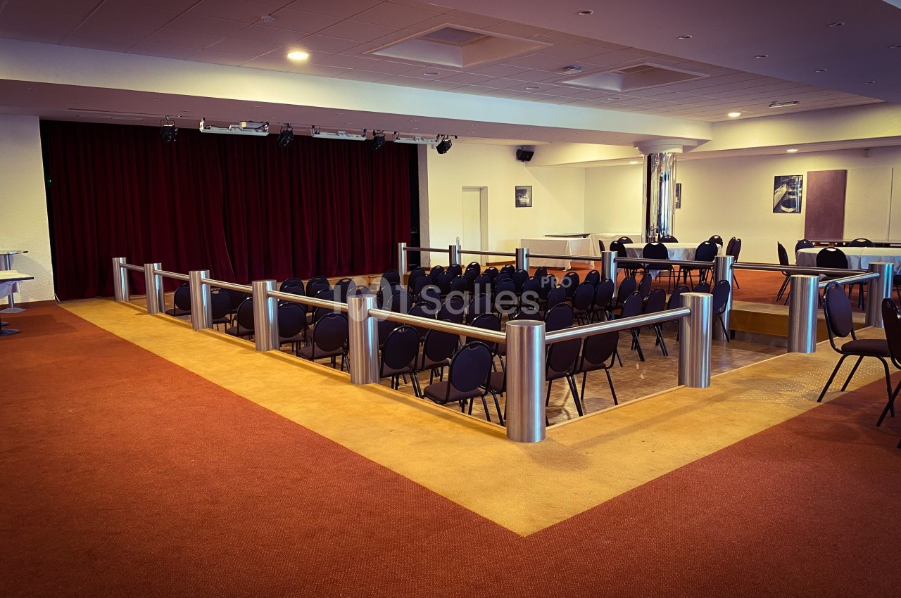 Salle de conférence vide avec des rangées de chaises noires, une scène avec rideaux rouges et un sol en moquette.