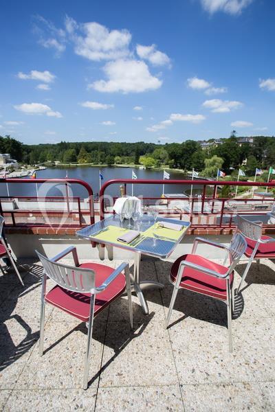 Table et chaises sur une terrasse ensoleillée avec vue sur un lac, des arbres et un ciel bleu parsemé de nuages.