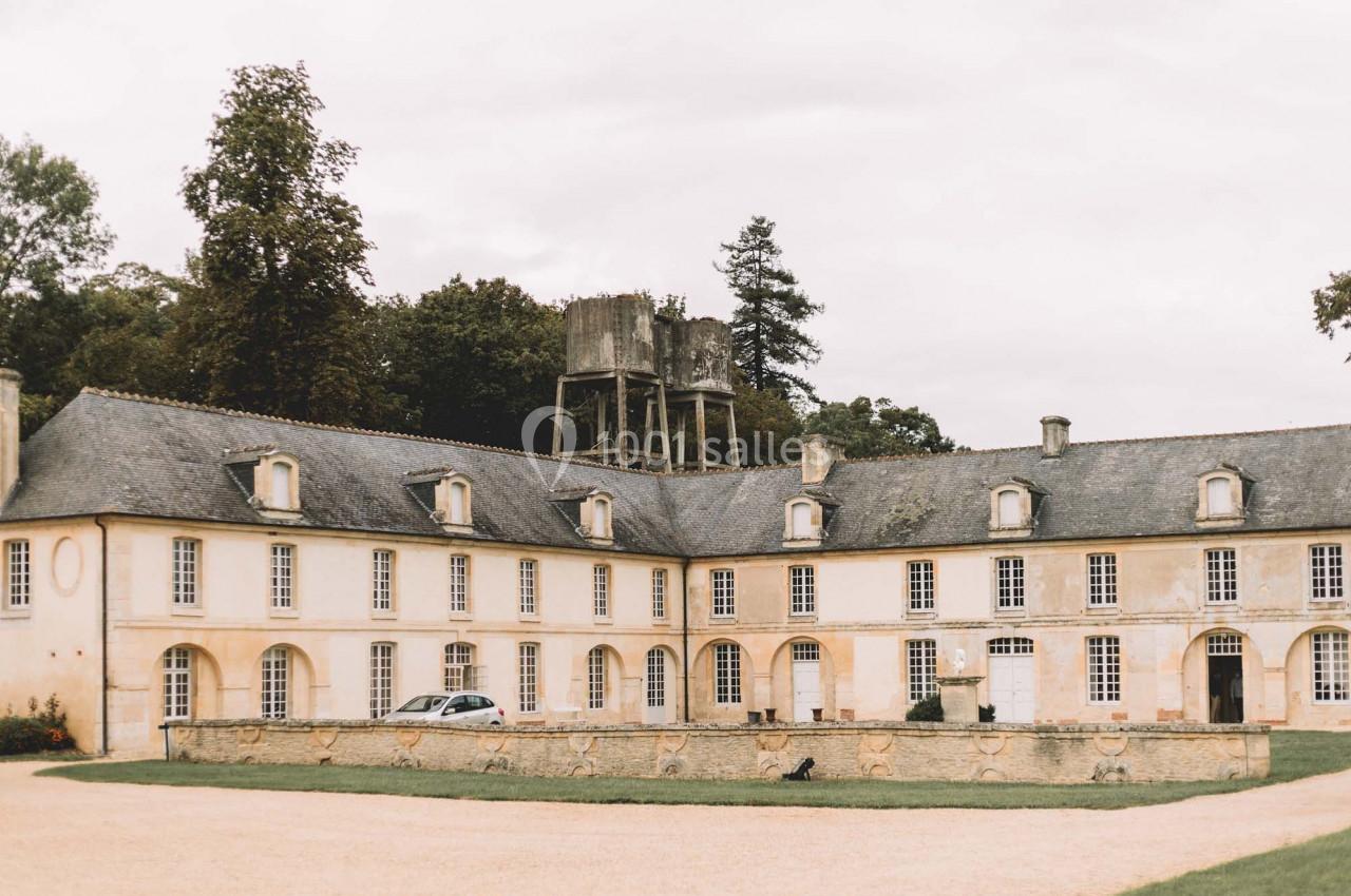 Bâtiment ancien en pierre avec toit en ardoise, cour centrale gravillonnée et structure en bois en arrière-plan.