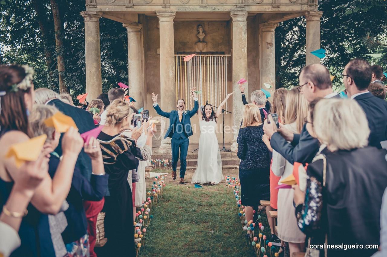 Un couple de mariés souriants marche entre des invités lançant des avions en papier dans un jardin devant un temple.