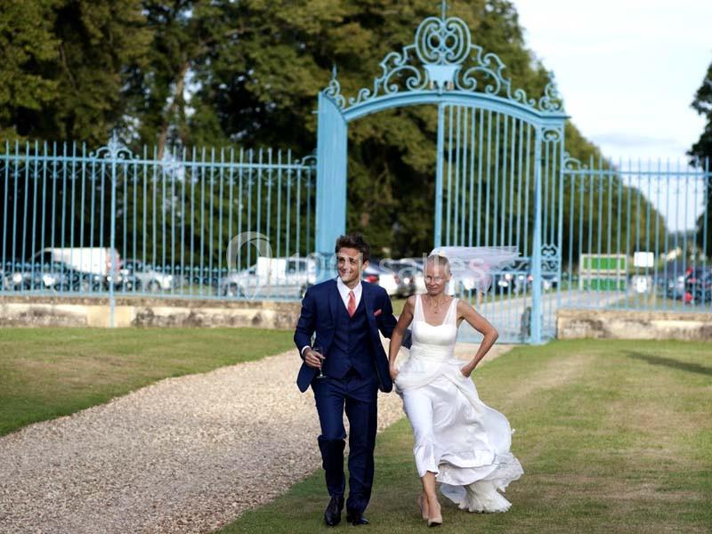 Un couple en tenue de mariage marche rapidement sur une allée gravillonnée devant une grille bleue dans un parc.