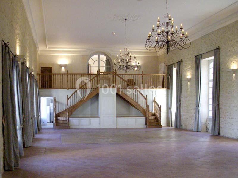 Grande salle lumineuse avec murs en pierre, lustres suspendus, mezzanine en bois et larges rideaux gris.