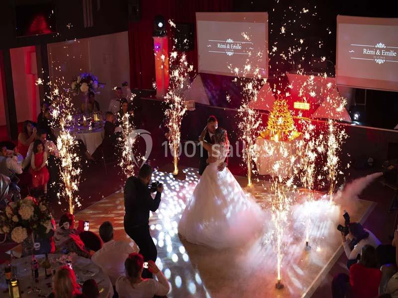 Un couple de mariés danse au centre d'une salle décorée, entouré de fontaines d'étincelles et de spectateurs assis.