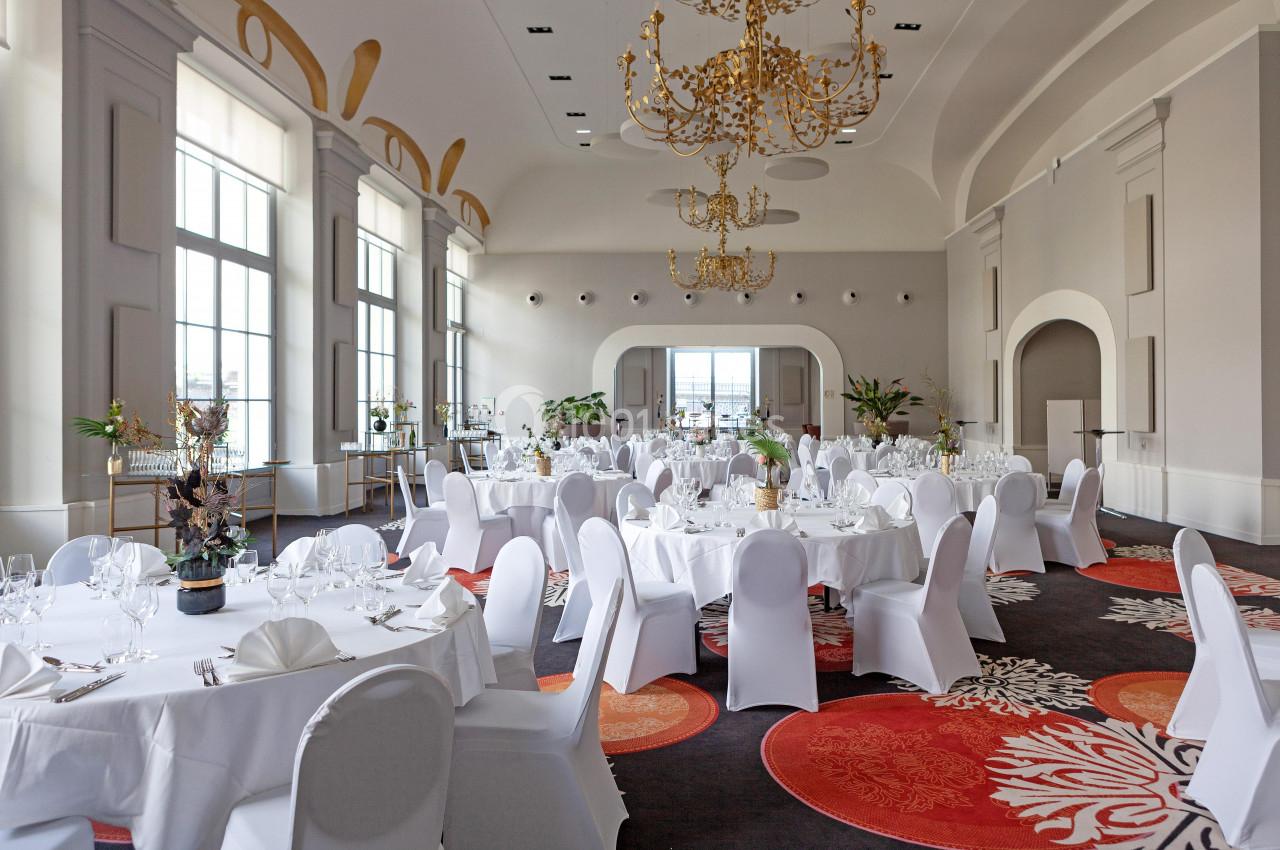 Salle de réception élégante avec tables rondes dressées, chaises blanches et lustres dorés sous un plafond voûté.