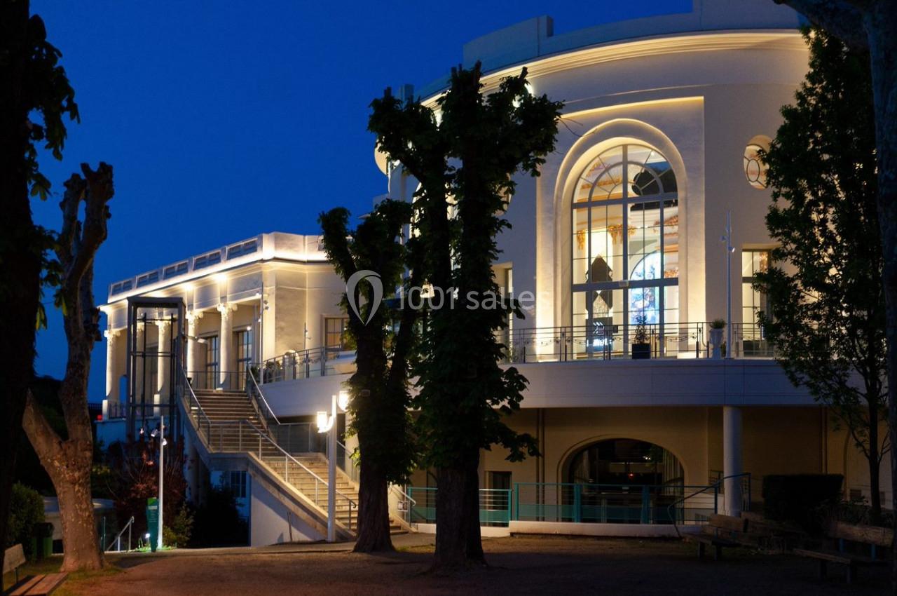 Façade illuminée d'un bâtiment élégant avec de grandes fenêtres et des arbres au premier plan, photographiée de nuit.
