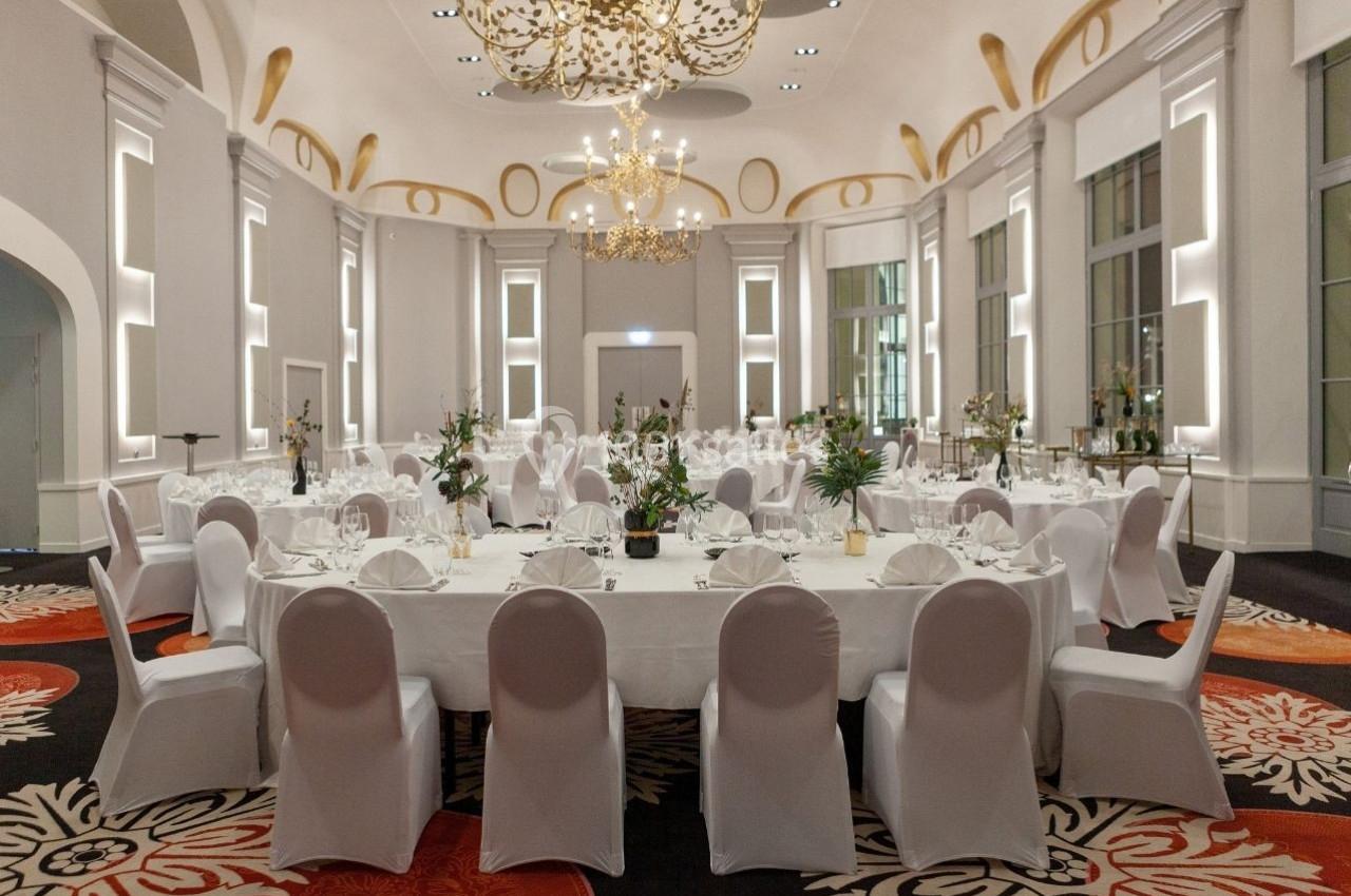 Salle de réception élégante avec tables rondes dressées, chaises blanches et lustres dorés suspendus.
