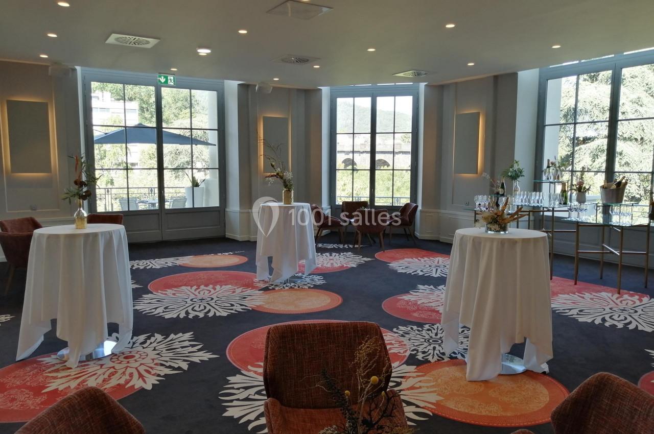Salle lumineuse avec tables hautes drapées de blanc, chaises, décor floral et grandes fenêtres donnant sur un paysage…