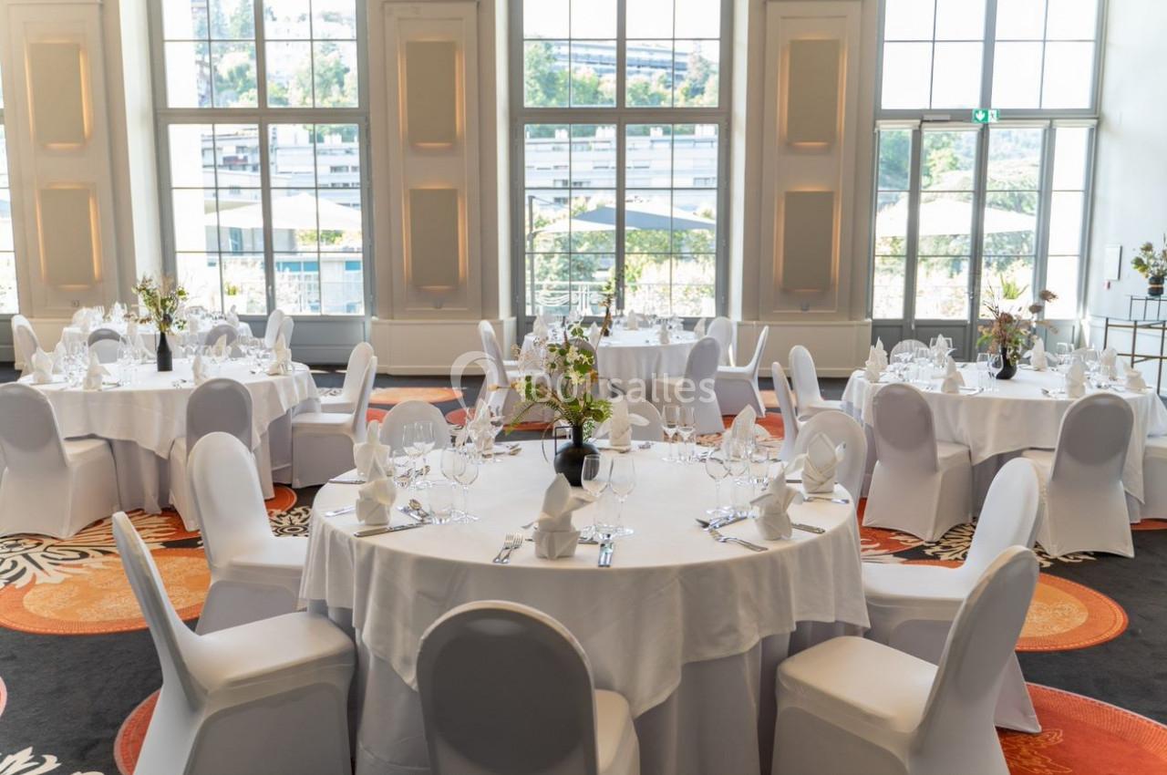 Salle lumineuse avec tables rondes dressées pour un événement, nappes blanches et chaises couvertes.