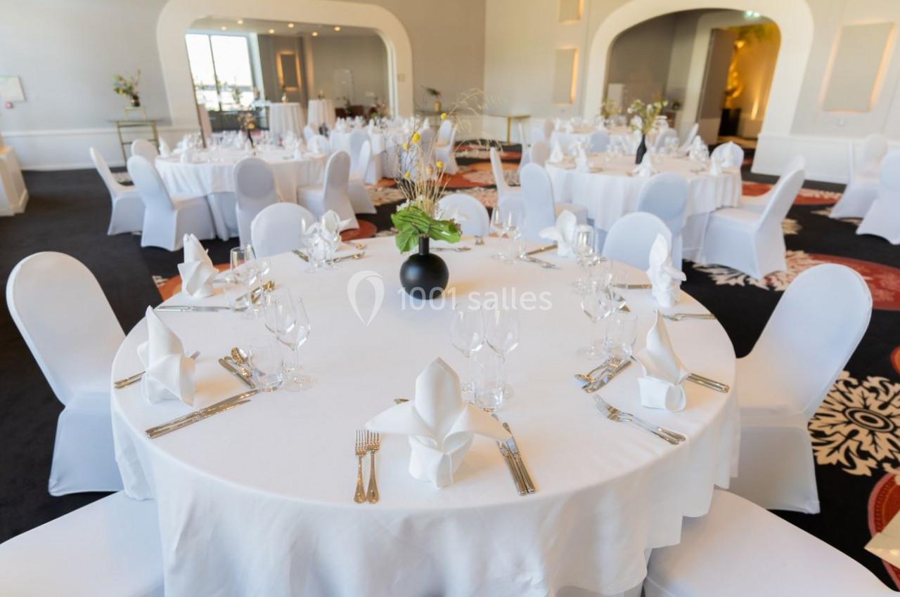 Salle de réception élégante avec des tables rondes dressées de nappes blanches, couverts et décorations florales.