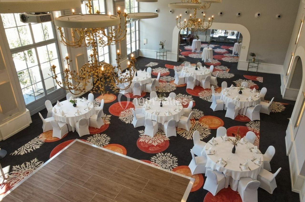 Salle de réception élégante avec tables rondes dressées, chaises blanches et lustres dorés, sur un tapis coloré.