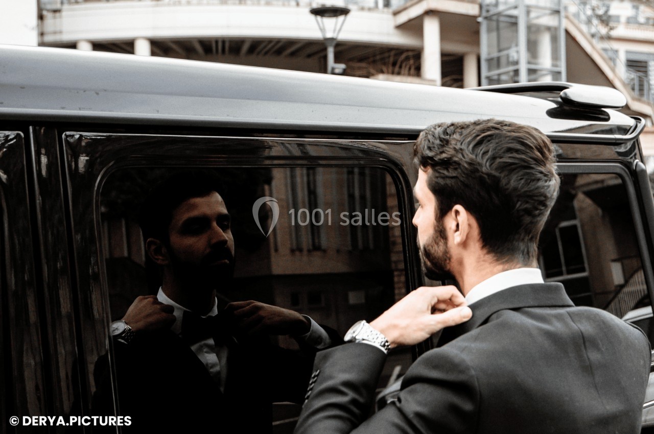 Un homme en costume ajuste son nœud papillon en se regardant dans la vitre d'un véhicule noir.