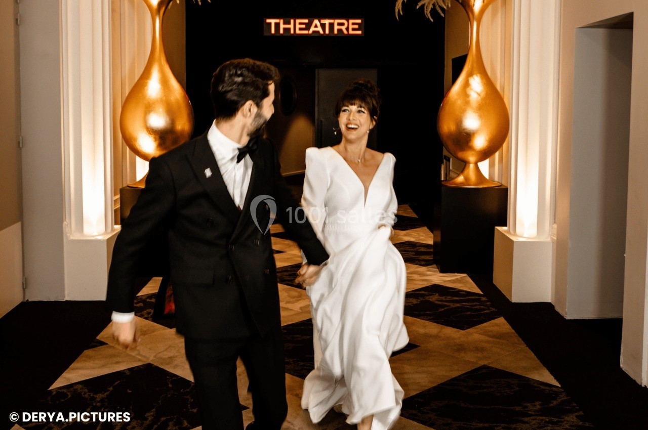 Un couple élégant souriant marche dans un couloir décoré de vases dorés, avec une enseigne ’Theatre’ en arrière-plan.
