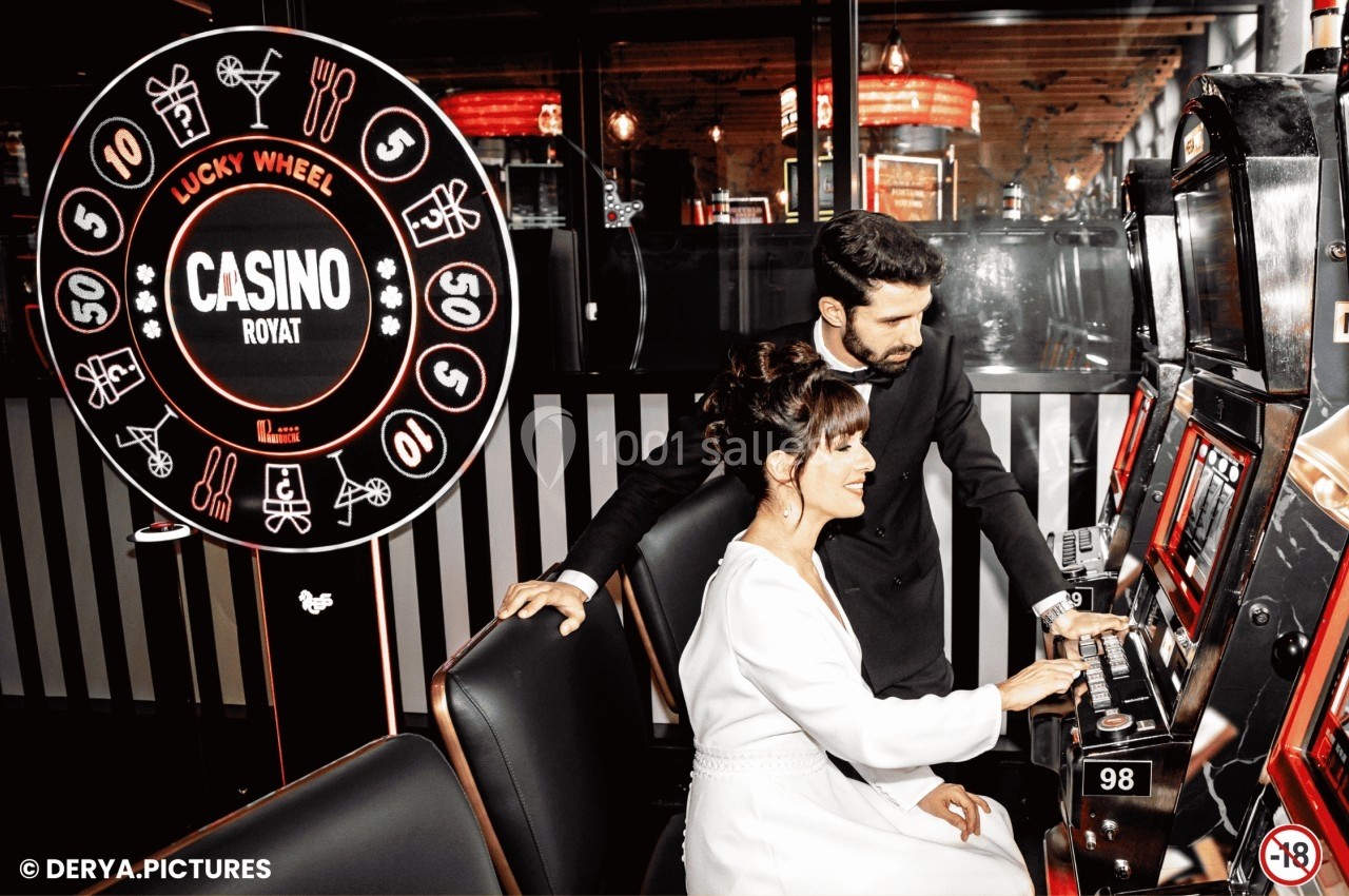 Un homme et une femme jouent à une machine à sous dans un casino, avec une roue de la chance en arrière-plan.