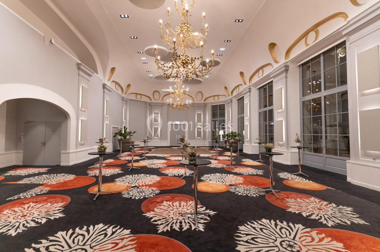 Salle élégante avec lustres dorés, grandes fenêtres, tapis coloré à motifs floraux et tables hautes dispersées.