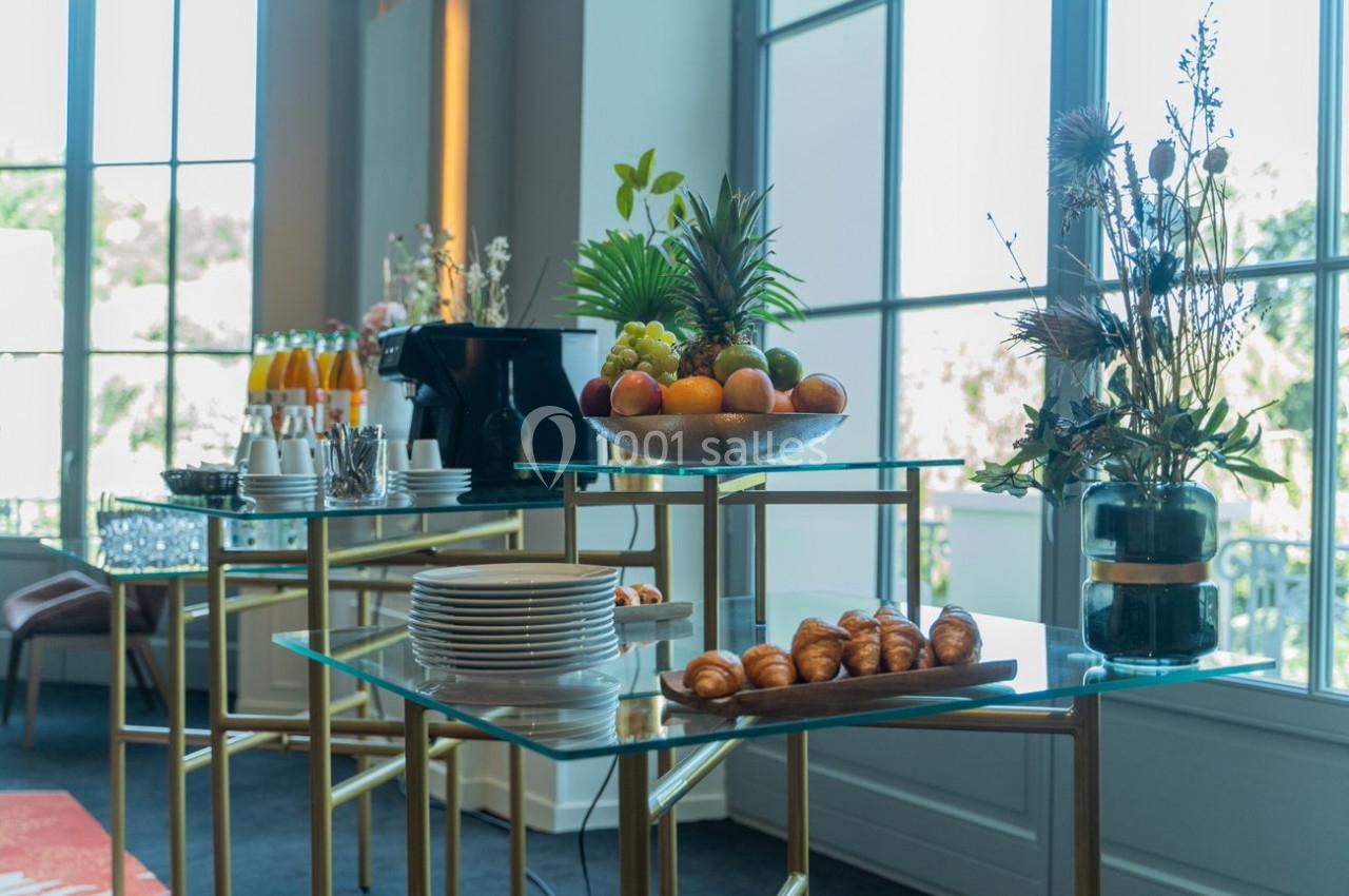 Buffet avec fruits, viennoiseries, boissons et vaisselle sur des tables en verre près de grandes fenêtres lumineuses.