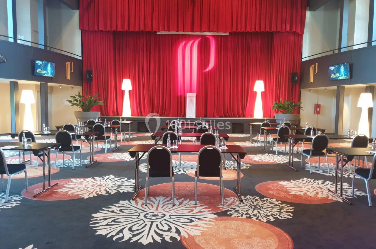 Salle de conférence avec des tables et chaises espacées, éclairée par des lampes et un rideau rouge en arrière-plan.