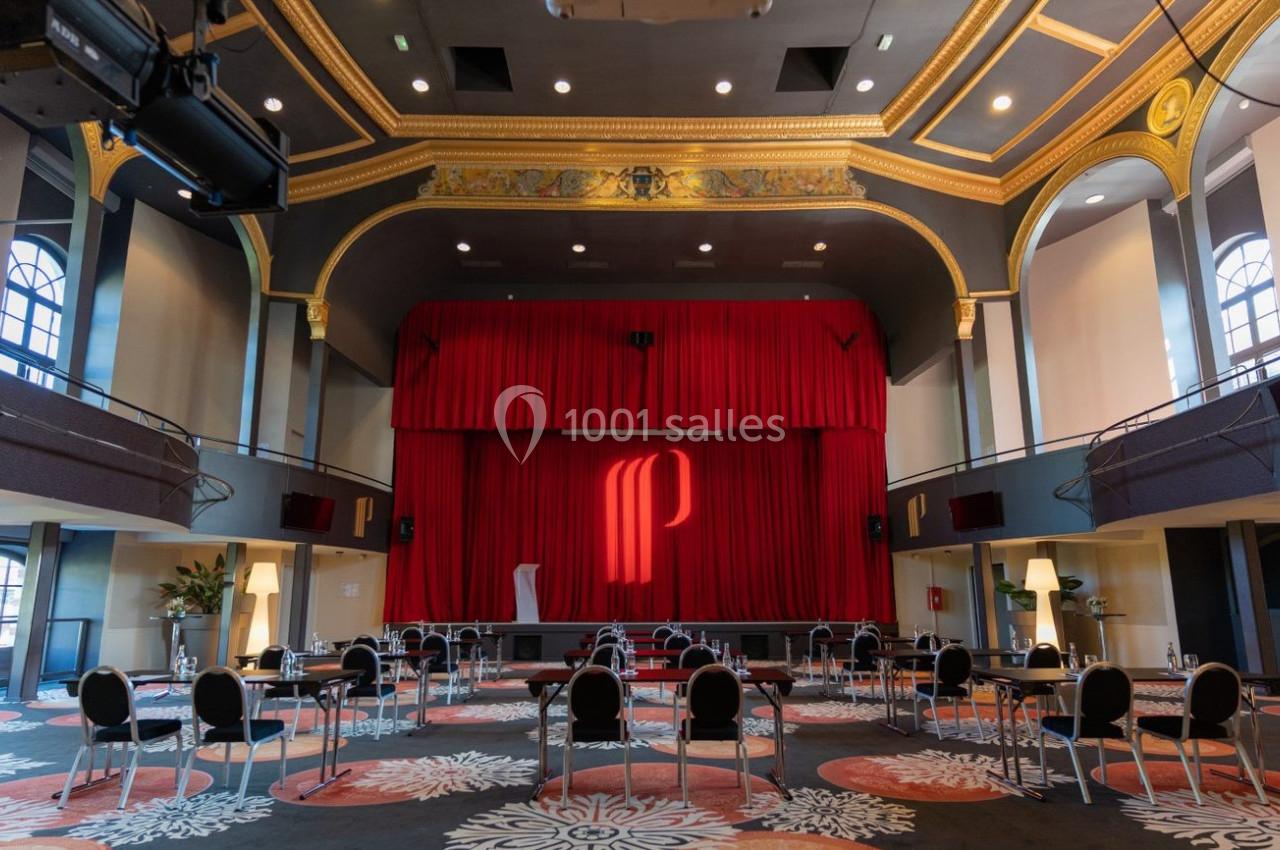 Salle de conférence élégante avec scène, rideaux rouges, logo lumineux et chaises disposées en rangées espacées.