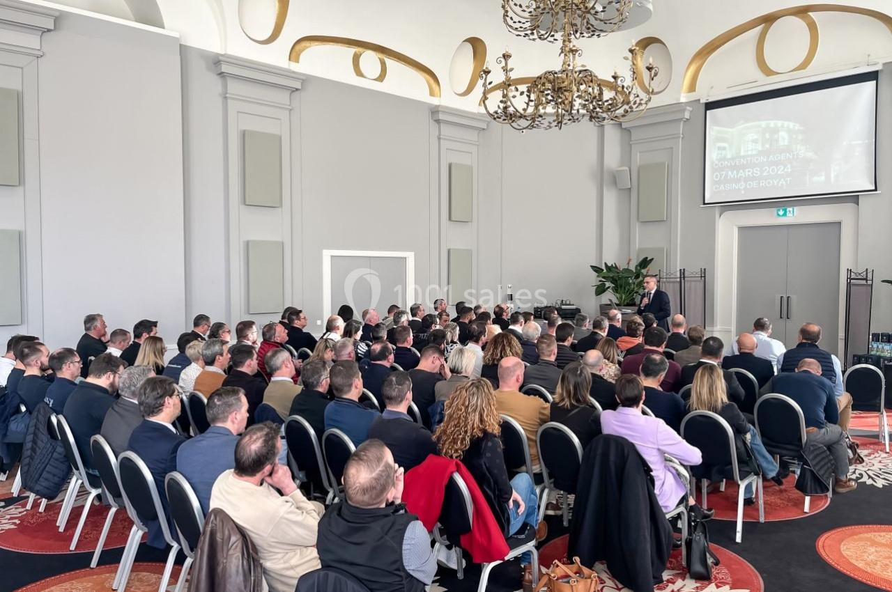 Un groupe de personnes assises dans une salle de conférence lumineuse, écoutant un orateur debout devant un écran.