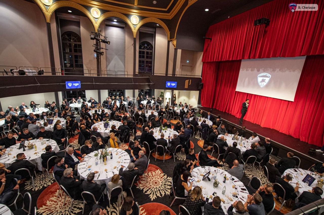 Salle de réception avec des tables rondes occupées par des convives, face à une scène avec un écran de projection.