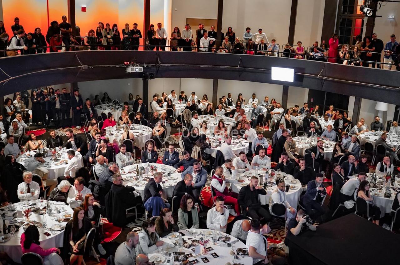 Salle comble avec des invités assis autour de tables rondes lors d'un événement, sous un éclairage chaleureux.