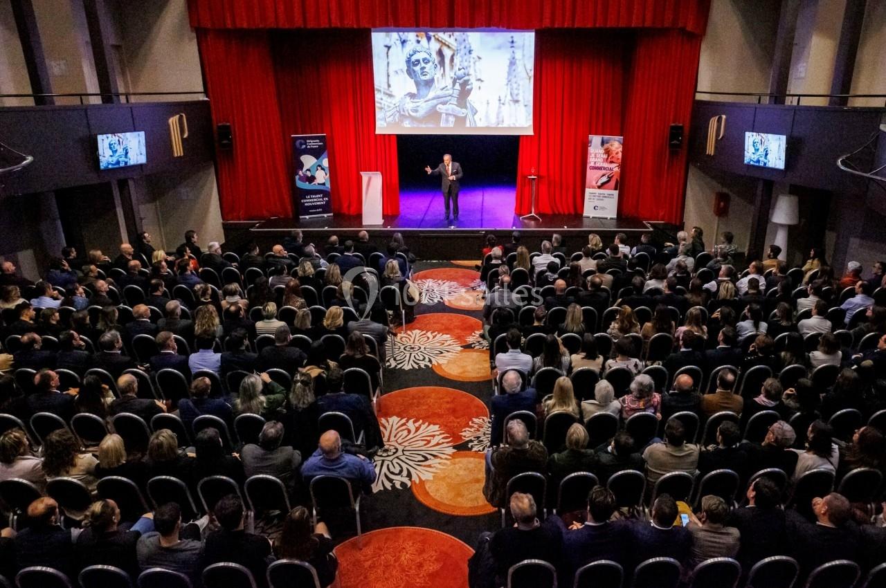 Salle de conférence avec un orateur sur scène devant un public, écran projetant une image artistique en arrière-plan.