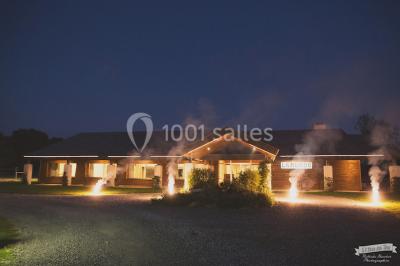 Vue d'une grange en bois entourée d'arbres, avec des bancs disposés en extérieur sur une pelouse.