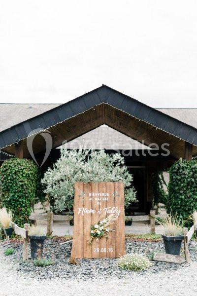 Entrée d'un bâtiment avec un panneau en bois décoré de fleurs, indiquant ’Bienvenue au mariage’ et une date.