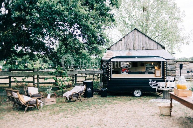 Camion de restauration noir stationné dans un jardin avec des chaises en bois, une grange et des arbres en arrière-plan.