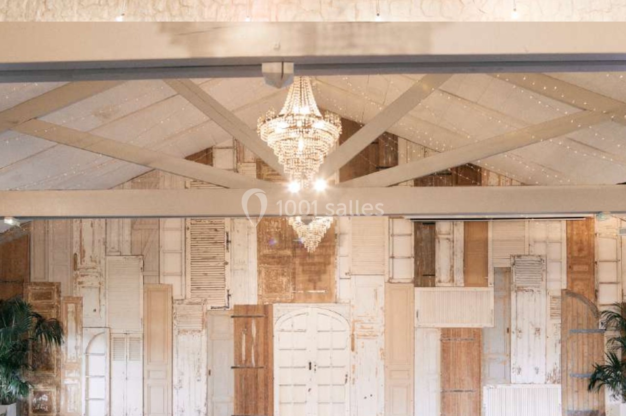 Mur décoratif composé de portes et volets anciens, éclairé par un lustre en cristal suspendu au plafond.