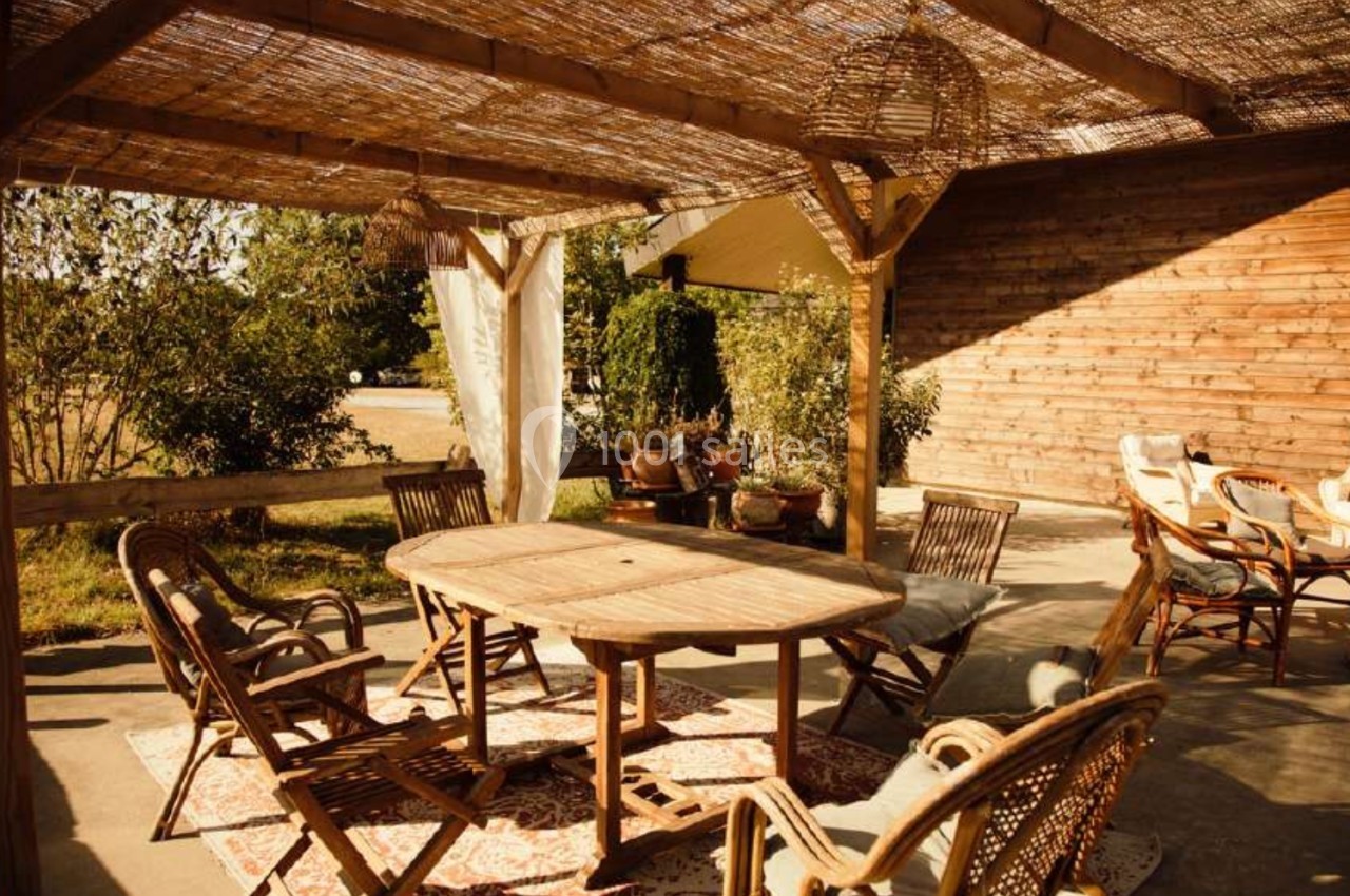 Terrasse en bois avec table et chaises sous une pergola en canisse, entourée de végétation et baignée de lumière naturelle.