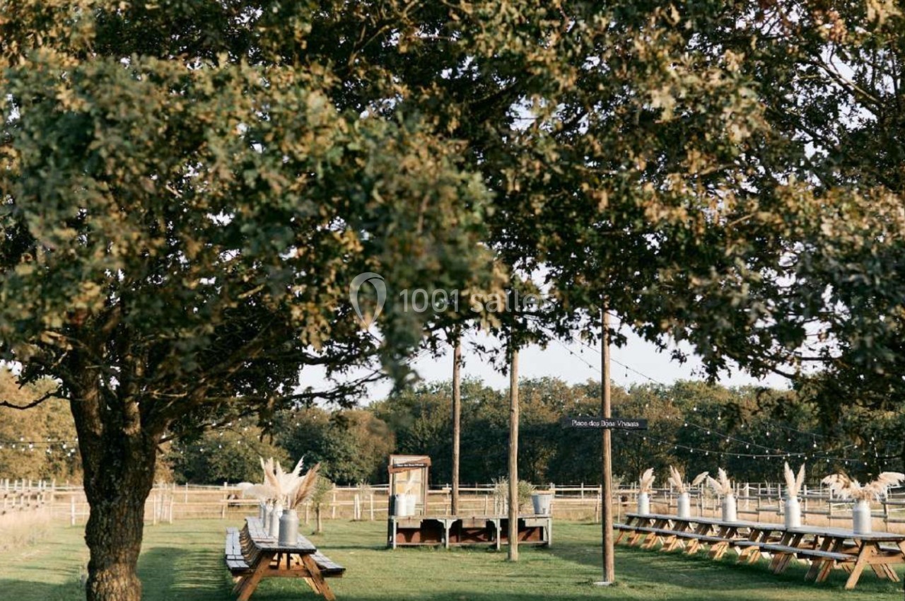 Tables en bois disposées en extérieur sous des arbres, avec des décorations et un panneau visible au centre.